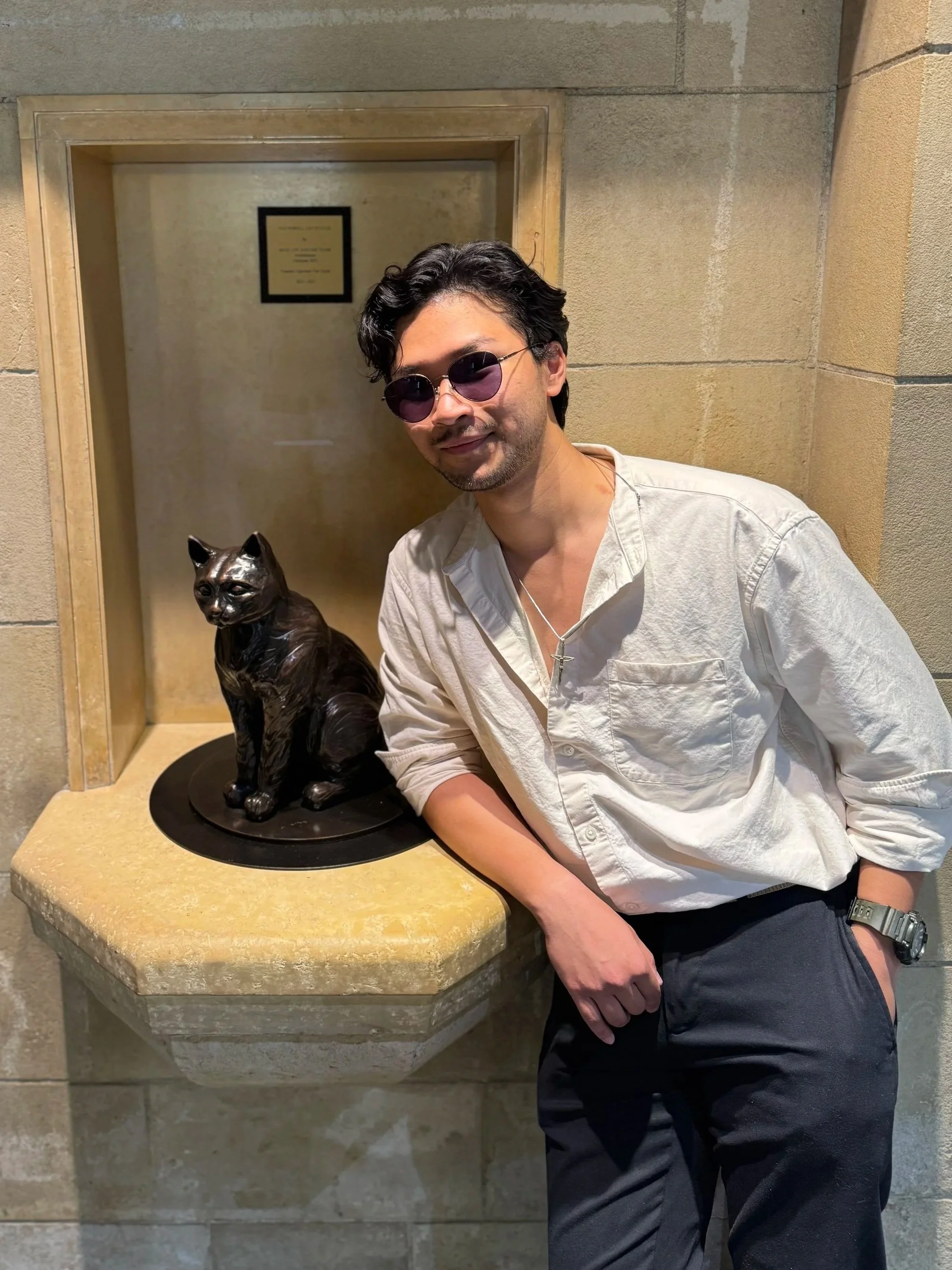A man in a white button-up shirt wearing sunglasses leaning next to a bronze cat statue adjacent to the wall.