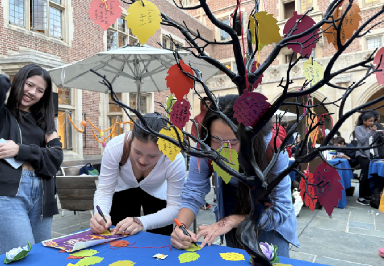 People participating in an outdoor fall celebration, decorating a table with colorful paper leaf cutouts and tree decorations in a courtyard.