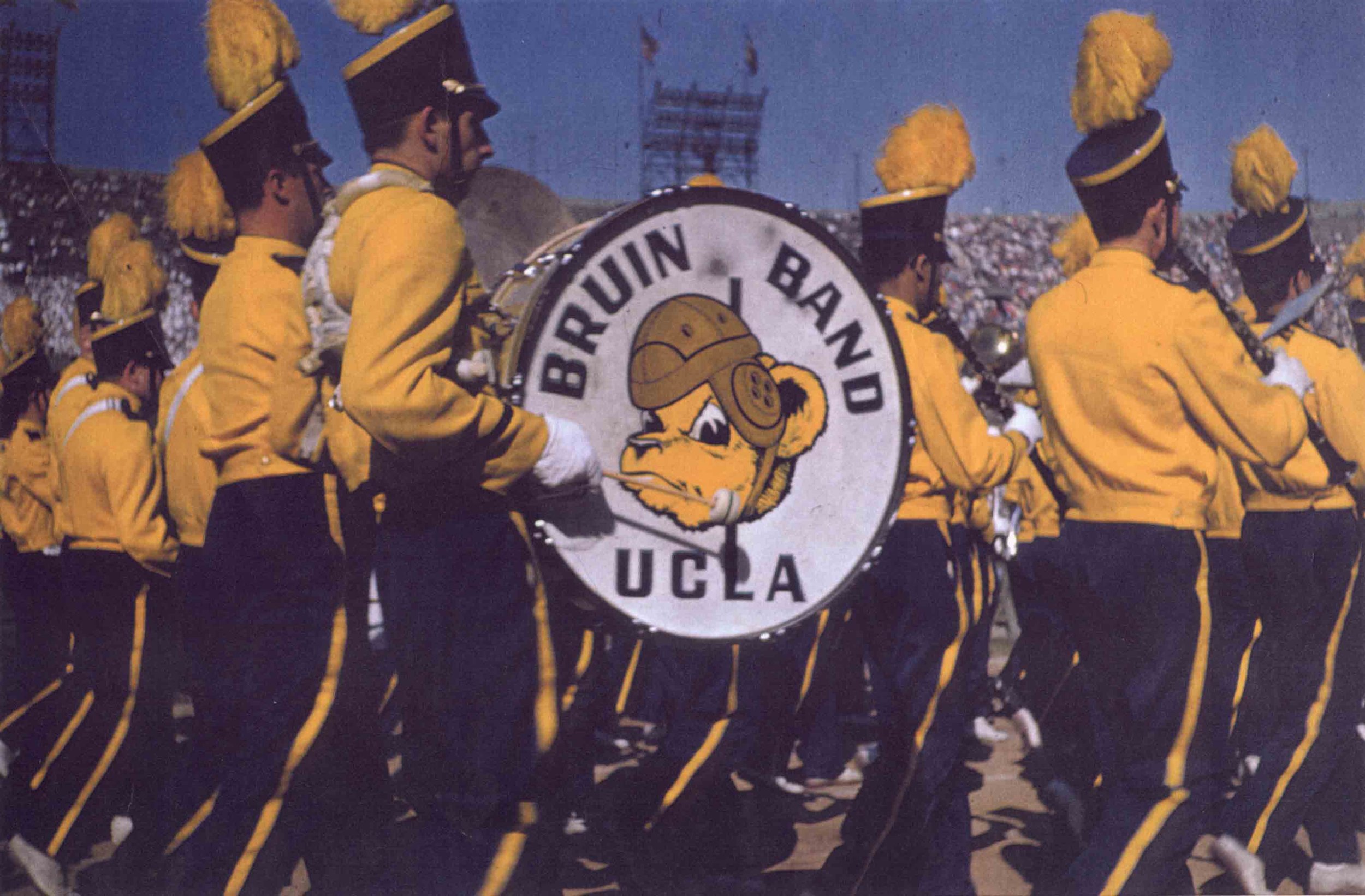 A vintage photograph of the UCLA marching band. At the center of the photo is a big bass drum with a drawing of Joe Bruin wearing a helmet and the words "Bruin Band UCLA". The band is wearing yellow and navy blue uniforms.