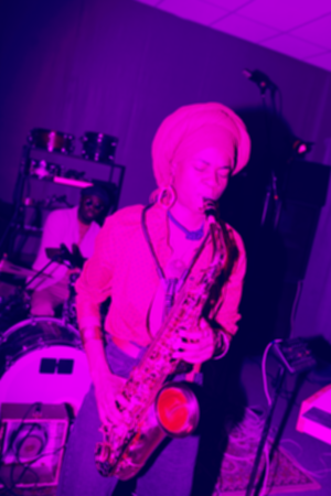 Musician plays a saxophone during an indoor live performance with a drummer in the background under purple stage lighting