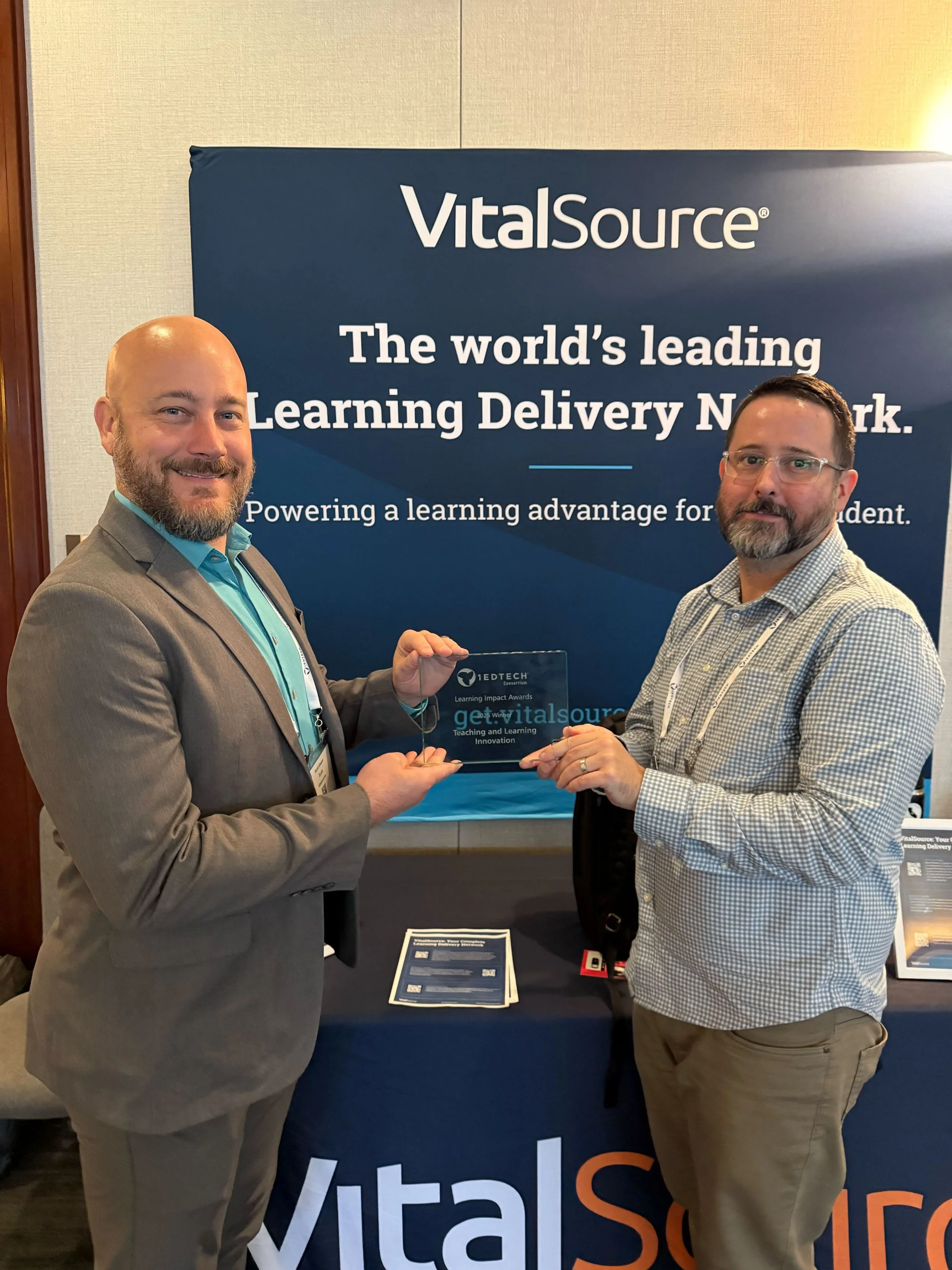 wo men in business attire hold a glass award at a VitalSource booth.