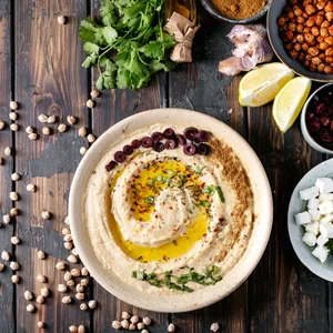 Overhead view of a bowl of hummus topped with olive oil, herbs, and olives, surrounded by fresh ingredients including chickpeas, garlic, lemon, cilantro, and spices on a wooden table