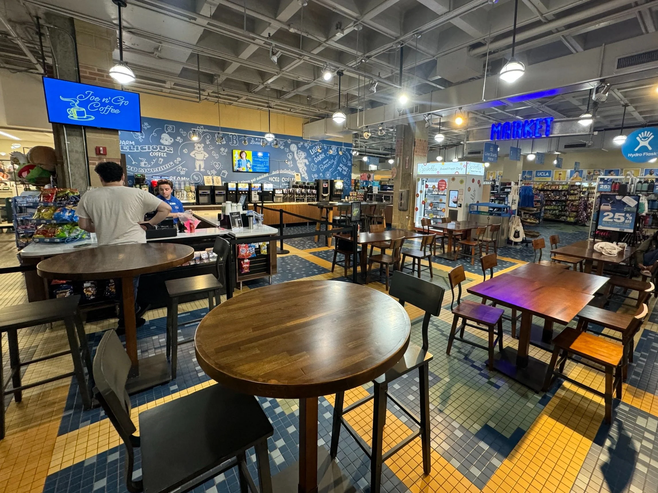 A brightly lit coffee space within a larger store featuring empty wooden tables and a cashier helping a customer.