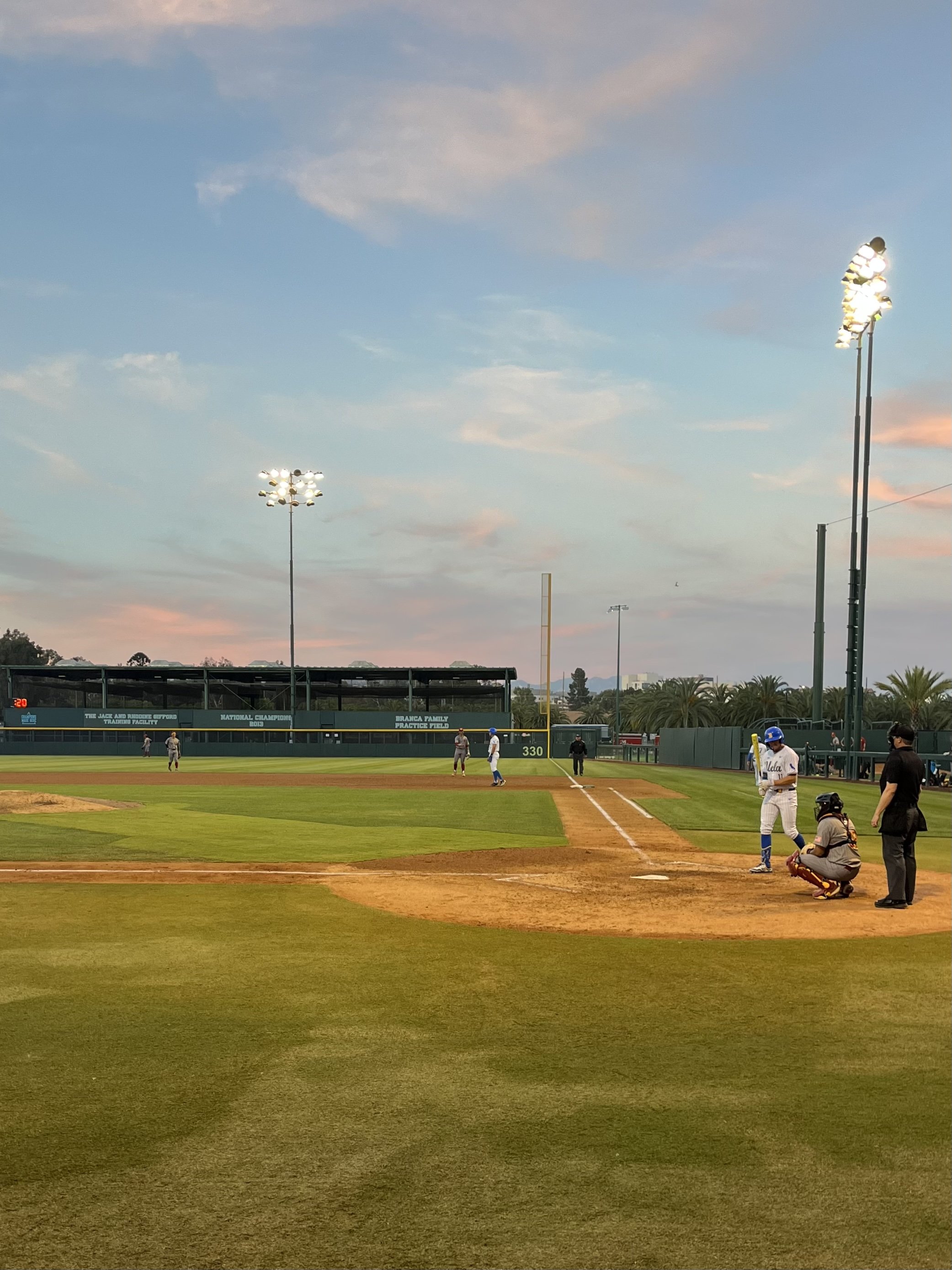 Man in white baseball uniform holding baseball bat walks towards home plate on baseball diamond with other players on the field