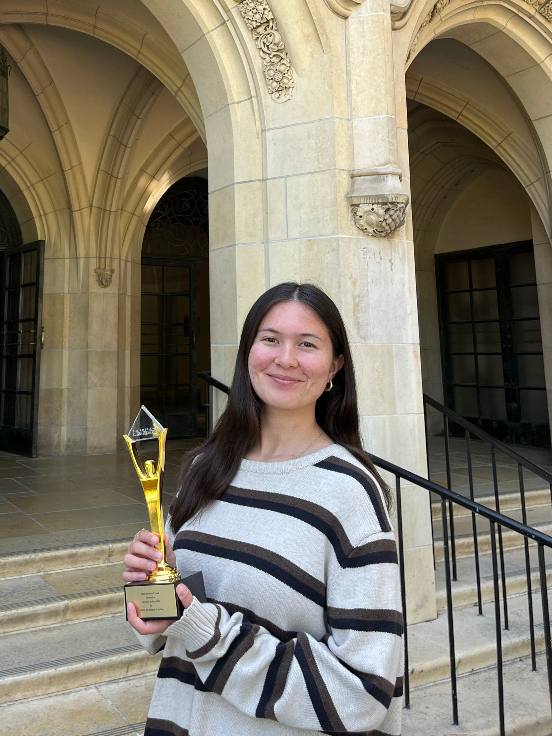 A woman in a striped sweater standing in front of stairs holding a gold award of a person lifting up a gold triangle with the text : "The American Business Awards."