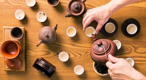 Overhead view of a traditional tea service with teapots, teacups, and hands pouring tea on a wooden table