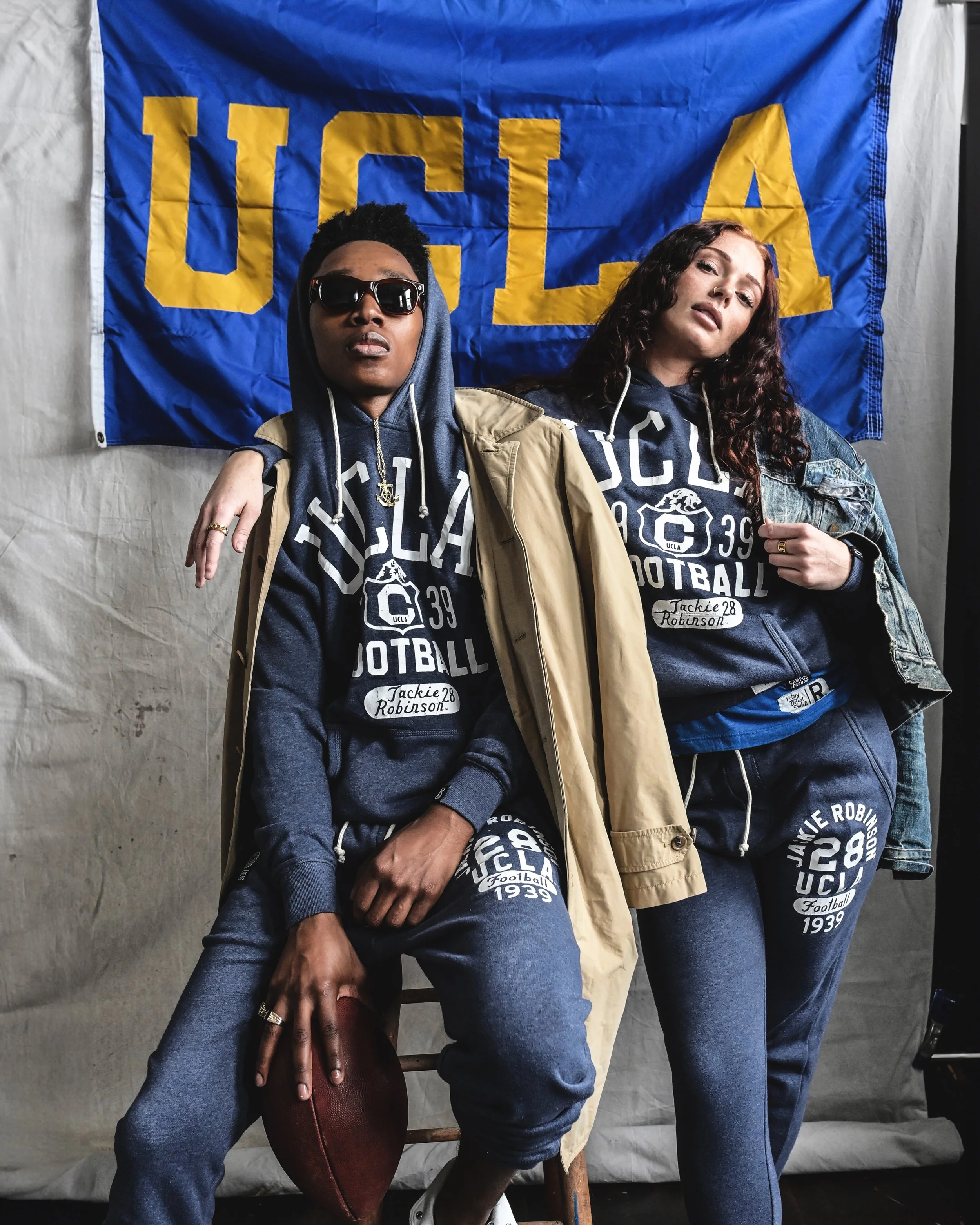 A man and woman wearing UCLA X Jackie Robinson navy track suits, in front of a blue UCLA banner with yellow text.