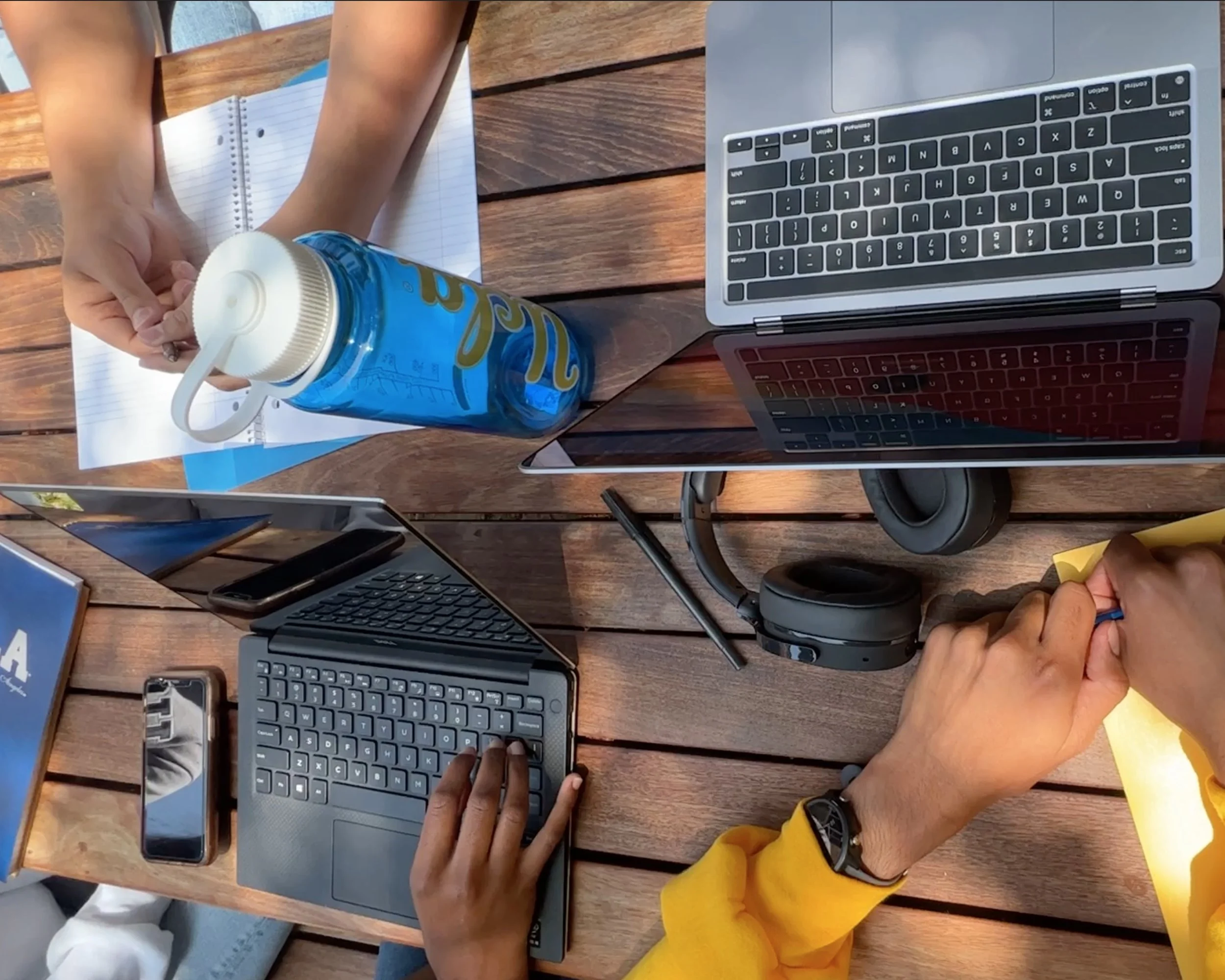 Bird's eye view of a tabletop. There are two computers, a water bottle, a phone, headphones, and some notebooks. There are three students working, but only their arms are visible.