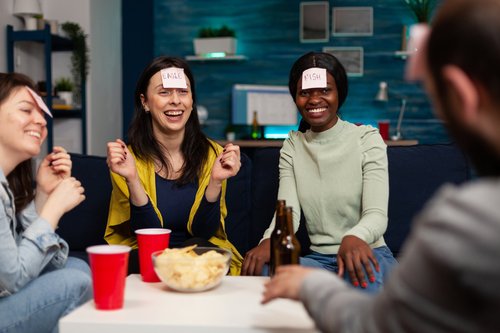A group of students laughing and playing a trivia game together, with clue cards on their foreheads, snacks, and drinks on the table