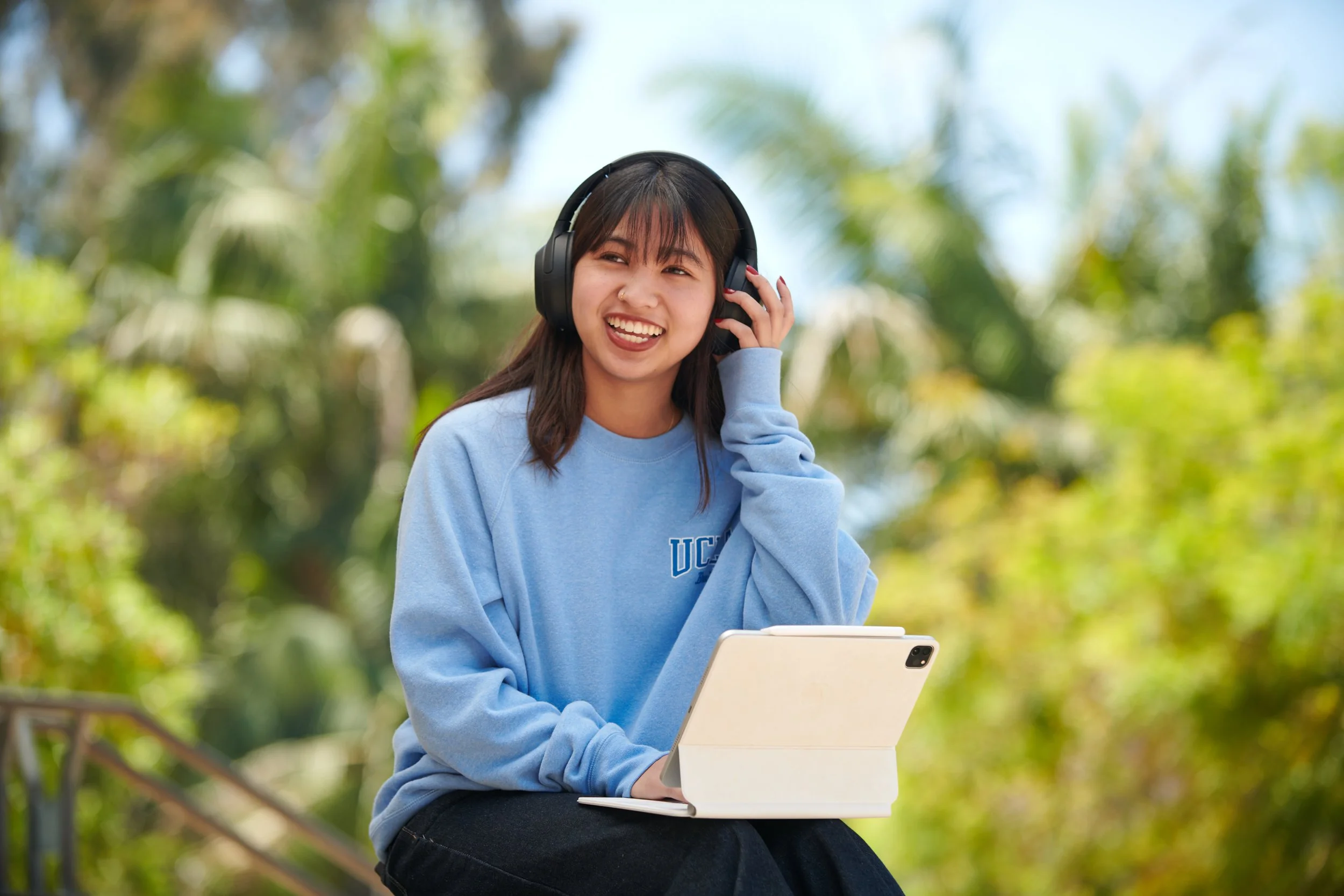Woman with headphones in UCLA blue crewneck holds iPad with white case