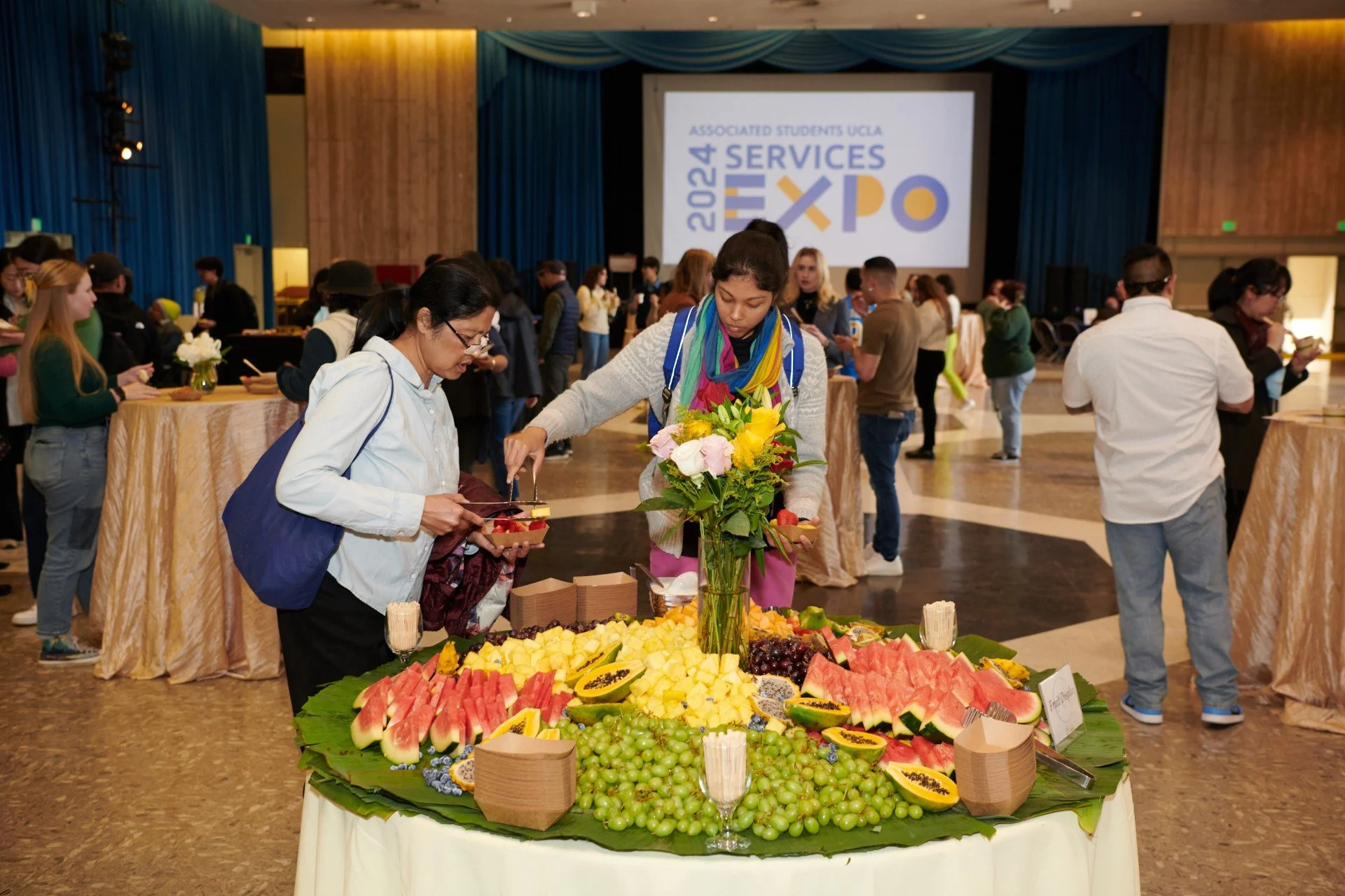 Two women serving fruit to their plate at a large ballroom gathering.