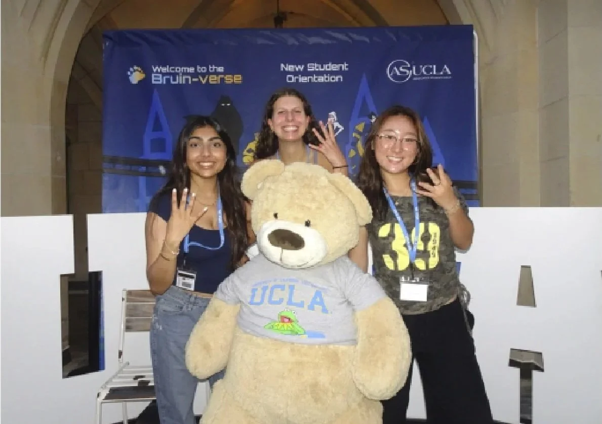 Three young women posing with a large teddy bear mascot at UCLA New Student Orientation, with a blue UCLA banner in the background.