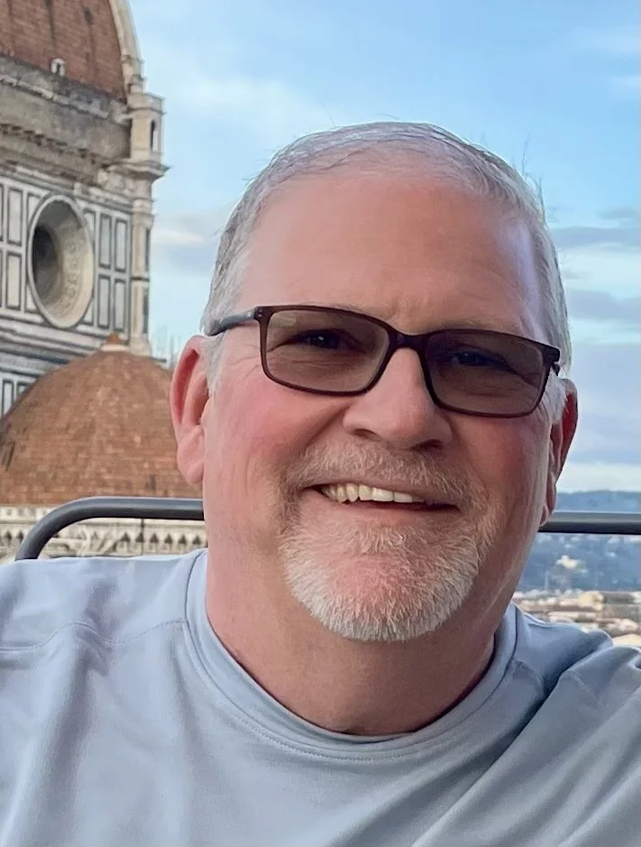A smiling man with grey hair, dark sunglasses wearing a light blue shirt.