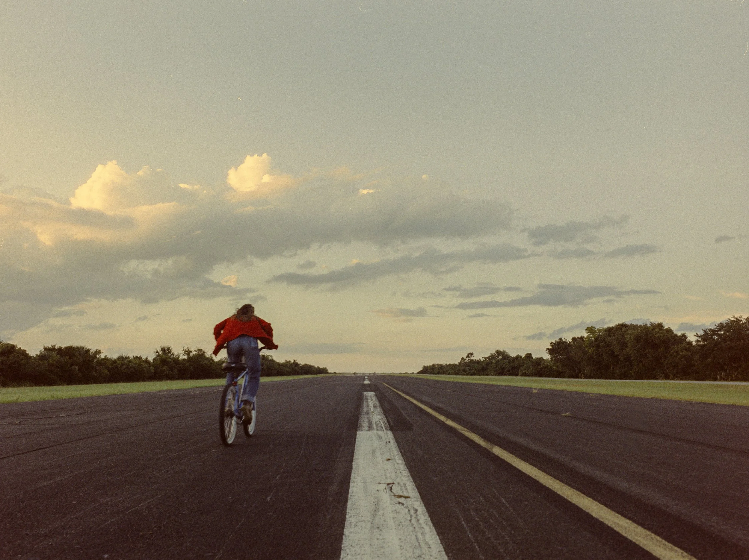 Person riding a bicycle down a long empty road in a rural area, with a cloudy sky overhead.