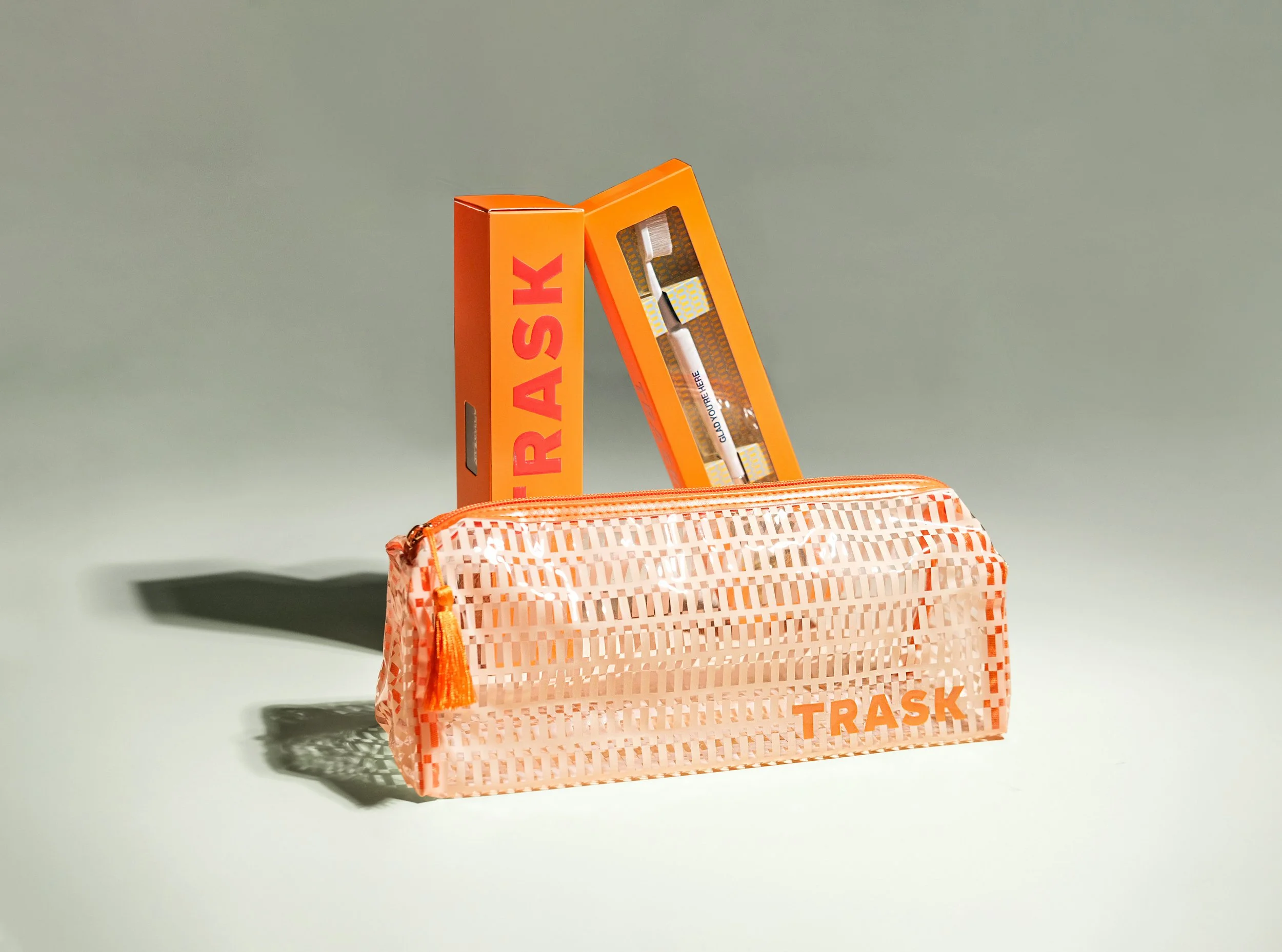 A pink and orange mesh trash bag with an orange zipper and orange tassel, and a box of toothbrushes with orange and yellow packaging, labeled "TRASH" and "GOT YOU COVERED".
