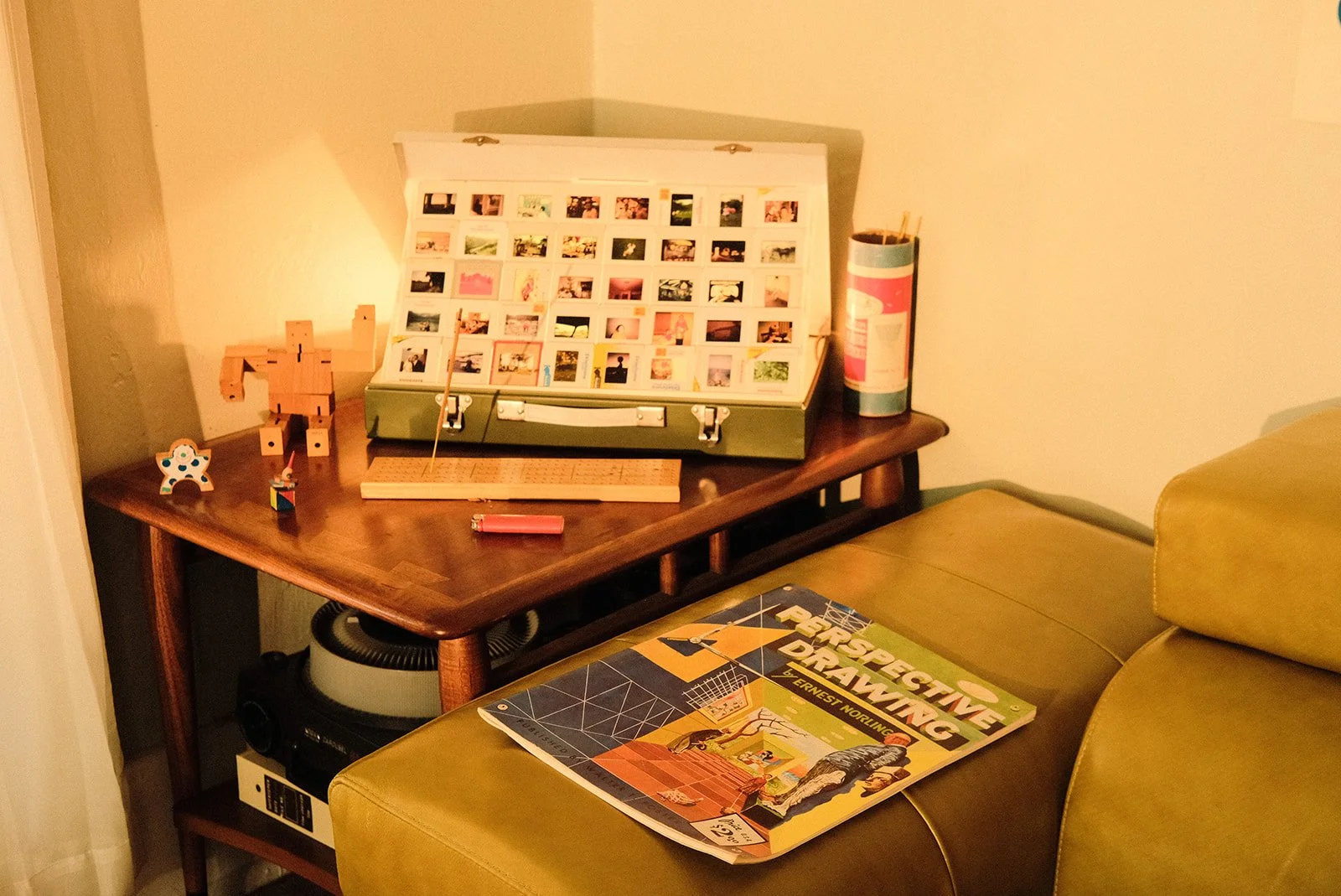 A wooden table with a white photo display, LEGO toy, small ceramic cat, and cylindrical container with colored markers. A green sofa with a book titled 'Perspective Drawing' on it. Part of a vacuum cleaner underneath the table and a yellow armchair.