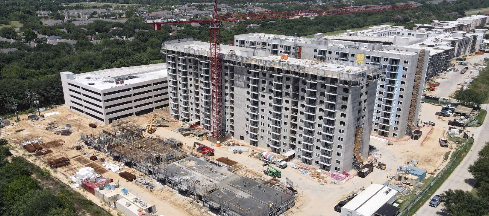 Aerial view of a large residential building under construction with a construction crane, construction materials, and vehicles on site, surrounded by green trees and nearby roads.