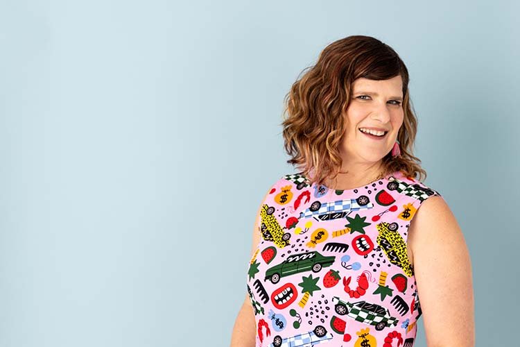 A woman with shoulder-length wavy brown hair, wearing a sleeveless top with colorful, playful patterns including fruits, cars, and abstract shapes, smiling and tilting her head slightly to the right against a light blue background.