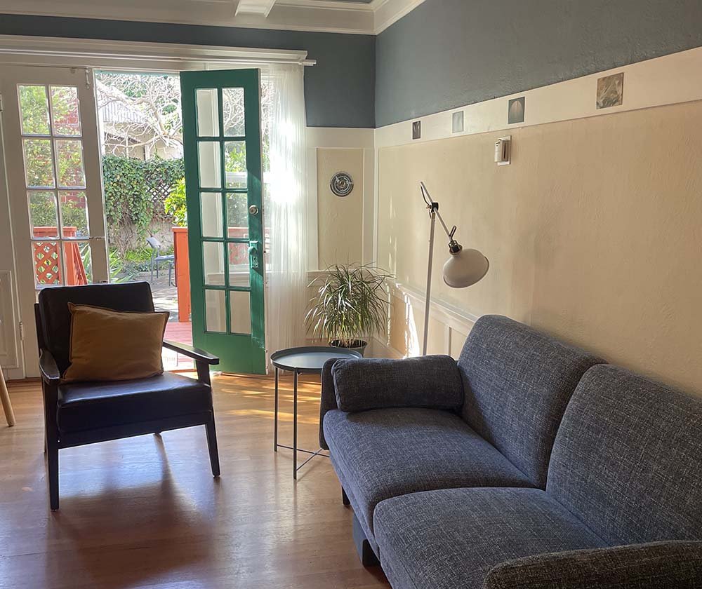 Living room with a gray sofa, black armchair with a yellow pillow, a small round table with a plant, a floor lamp, and an open door leading to a backyard with red deck and garden outside.