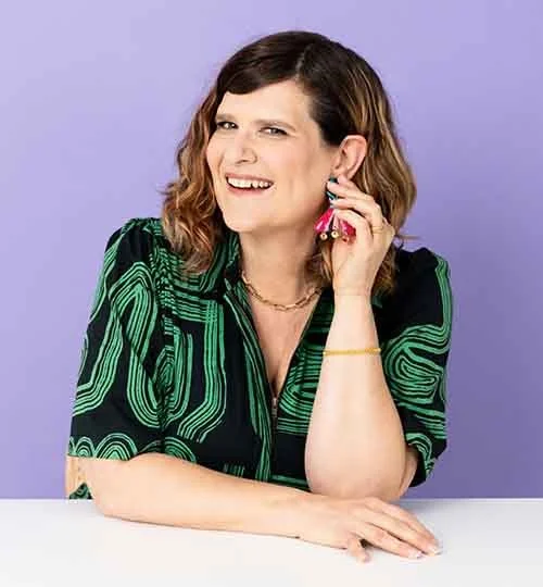 A woman with shoulder-length brown hair wearing a black and green patterned shirt, holding a pink and yellow toy in her right hand against a purple background.