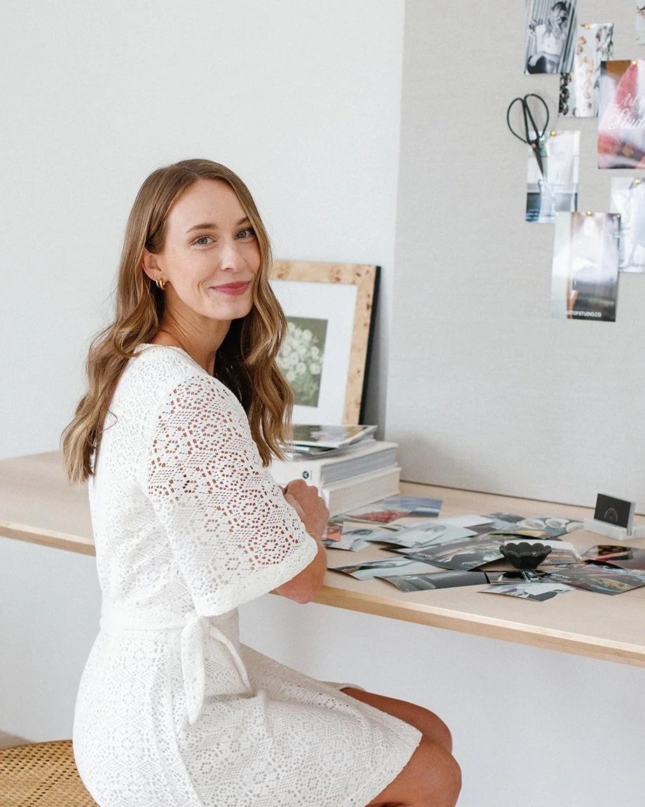 A woman with long wavy brown hair and light skin, wearing a white lace dress, sitting at a white table in an art studio or workspace, smiling at the camera. The table has various photographs, papers, and an ashtray on it. Behind her is a white board with photos clipped, and a framed artwork leaning against the wall.