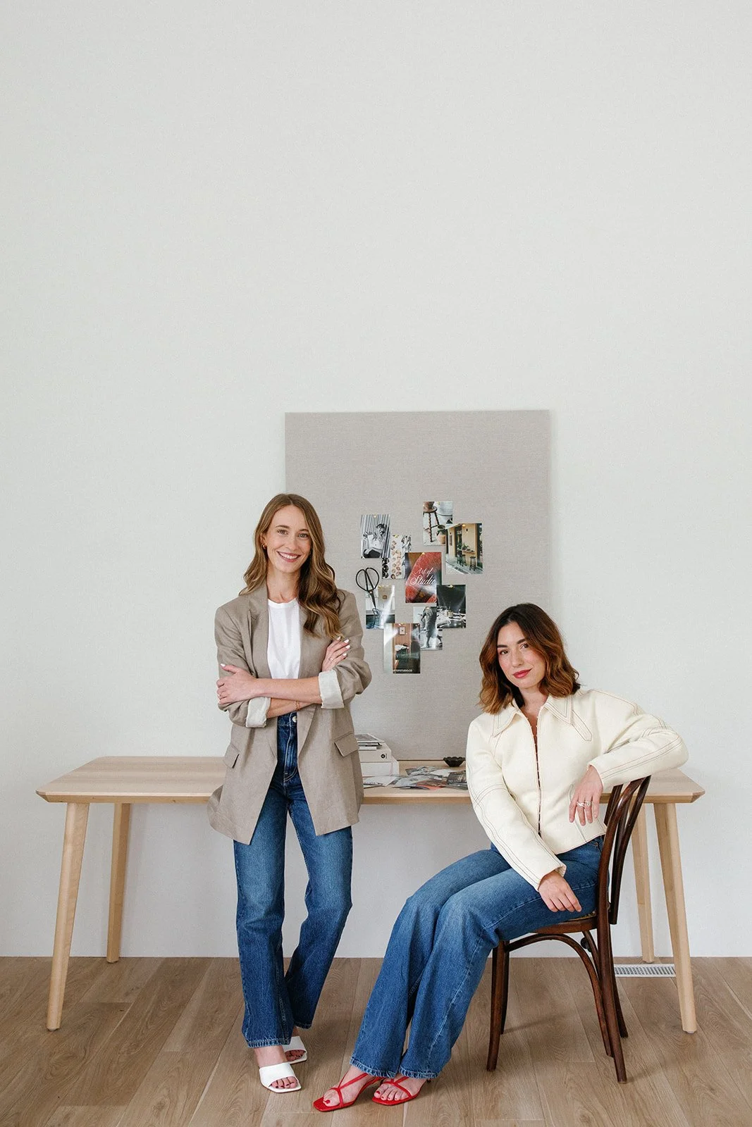 Two women in an office, one standing with arms crossed and the other sitting, smiling and looking at the camera, with a corkboard and photos on the wall behind them.