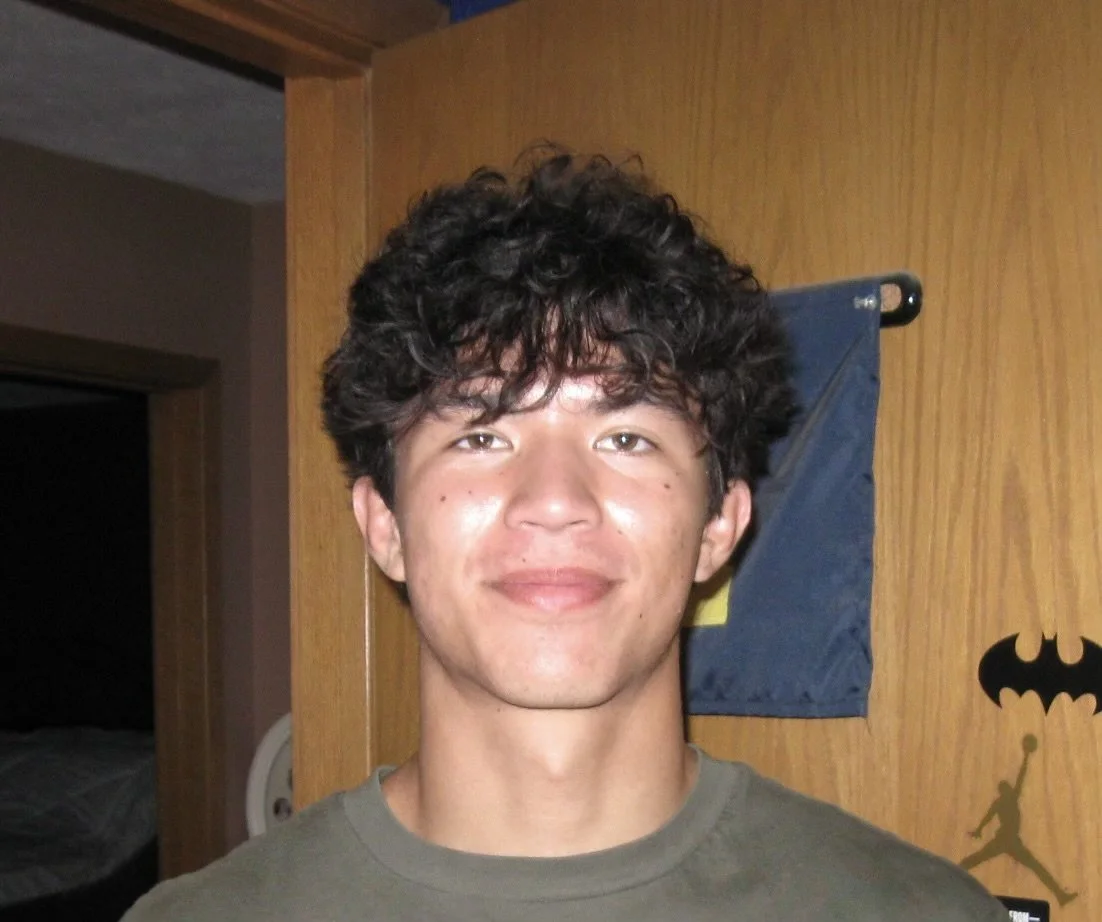 A person with curly dark hair smiling in front of a wooden door, with a Batman wall decal and a blue and yellow flag in the background.