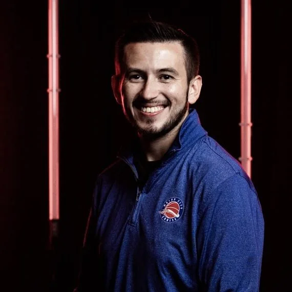 Smiling man in blue jacket with red and blue logo, standing against a dark background with red neon lights.