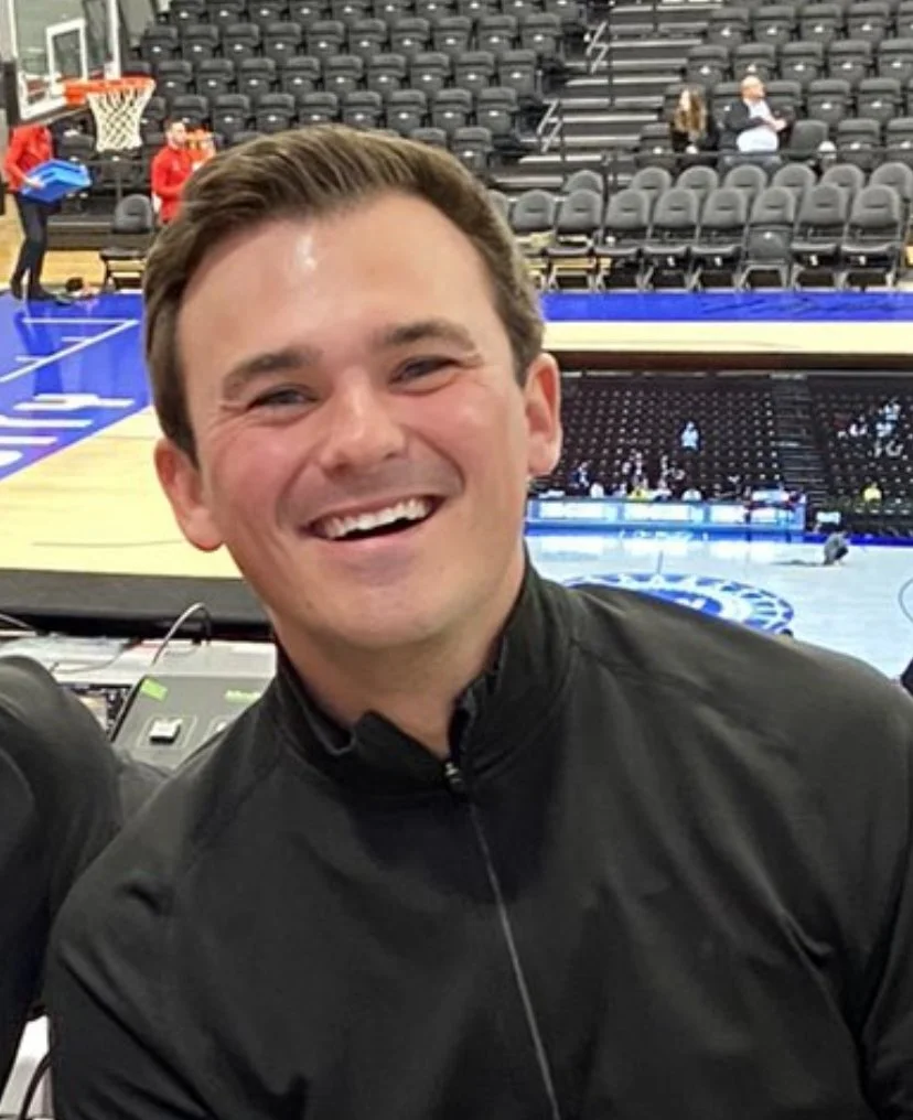 Person smiling in an indoor sports arena with basketball court and empty seats.