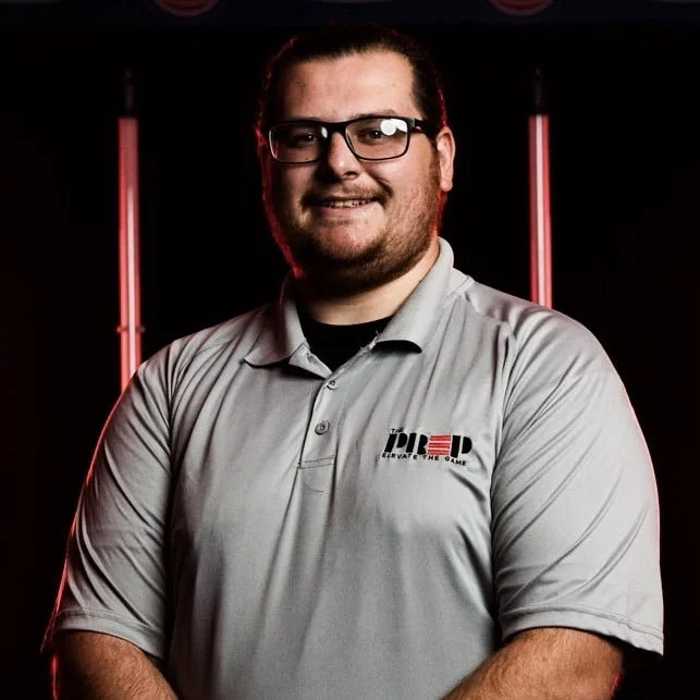 Person smiling in glasses and gray polo with logo, standing against black and red background.