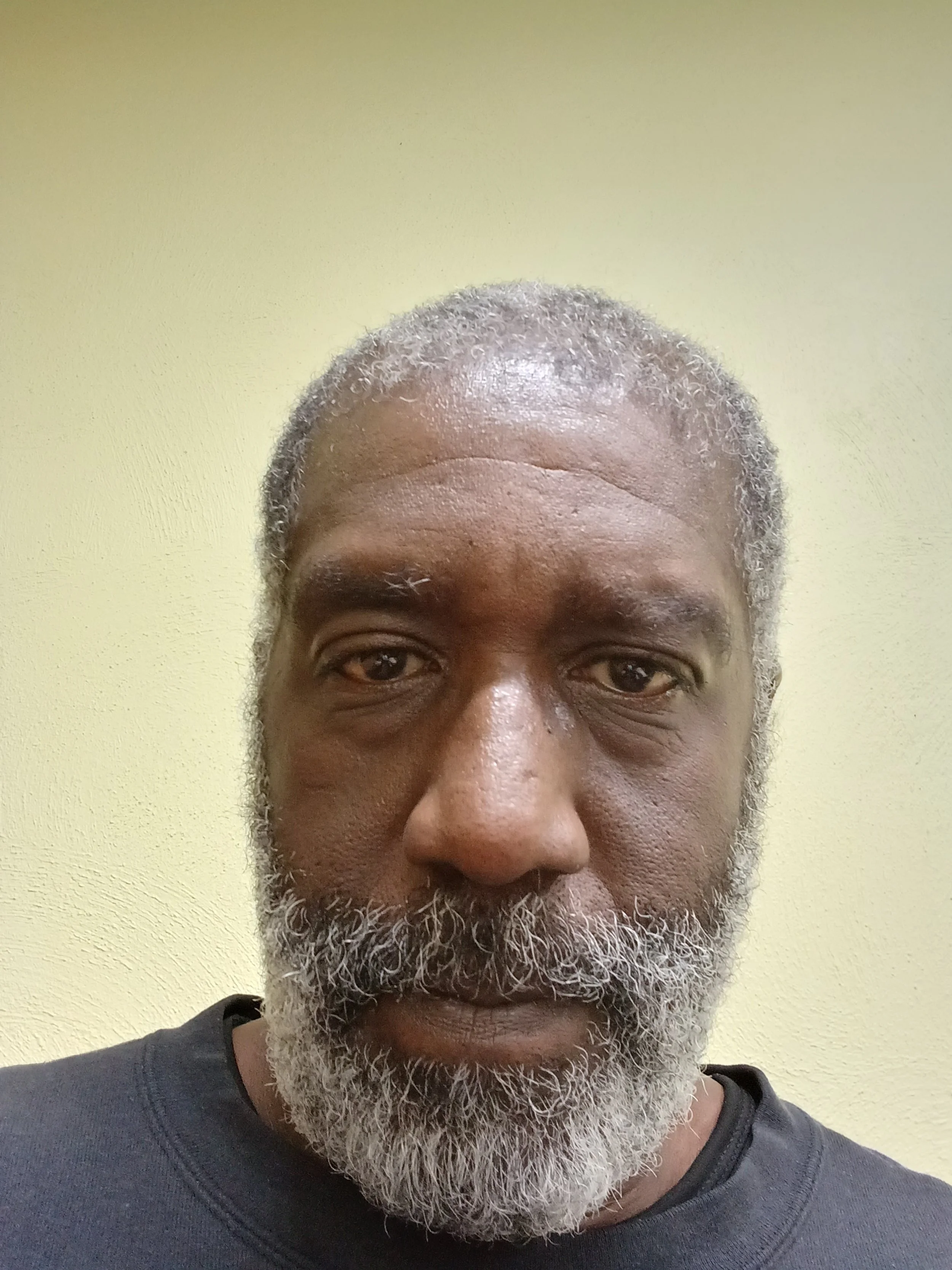 Close-up of a person with short hair and a beard, wearing a dark shirt, in front of a light-colored wall.