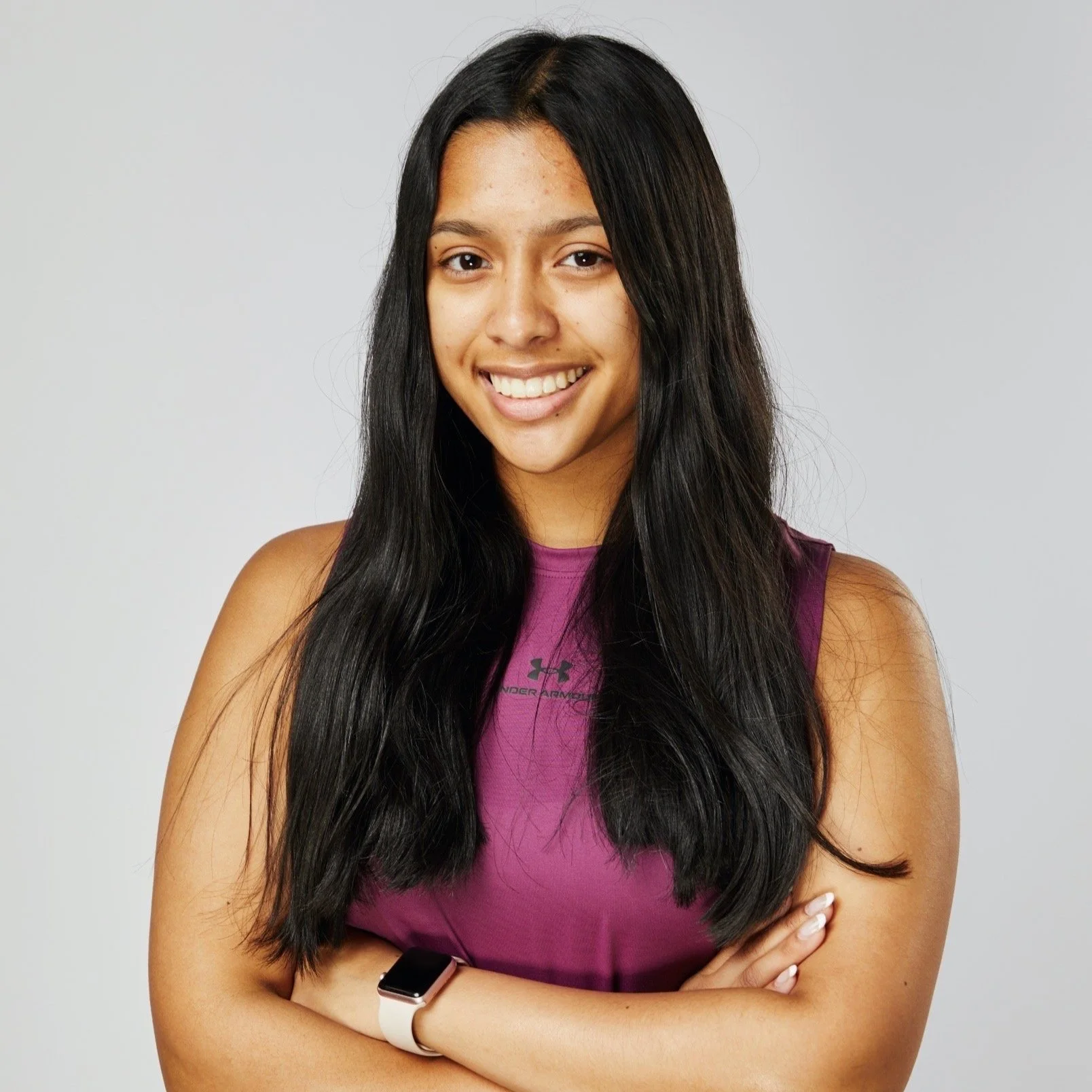 Smiling woman with long dark hair, wearing a purple tank top and smart watch, arms crossed.