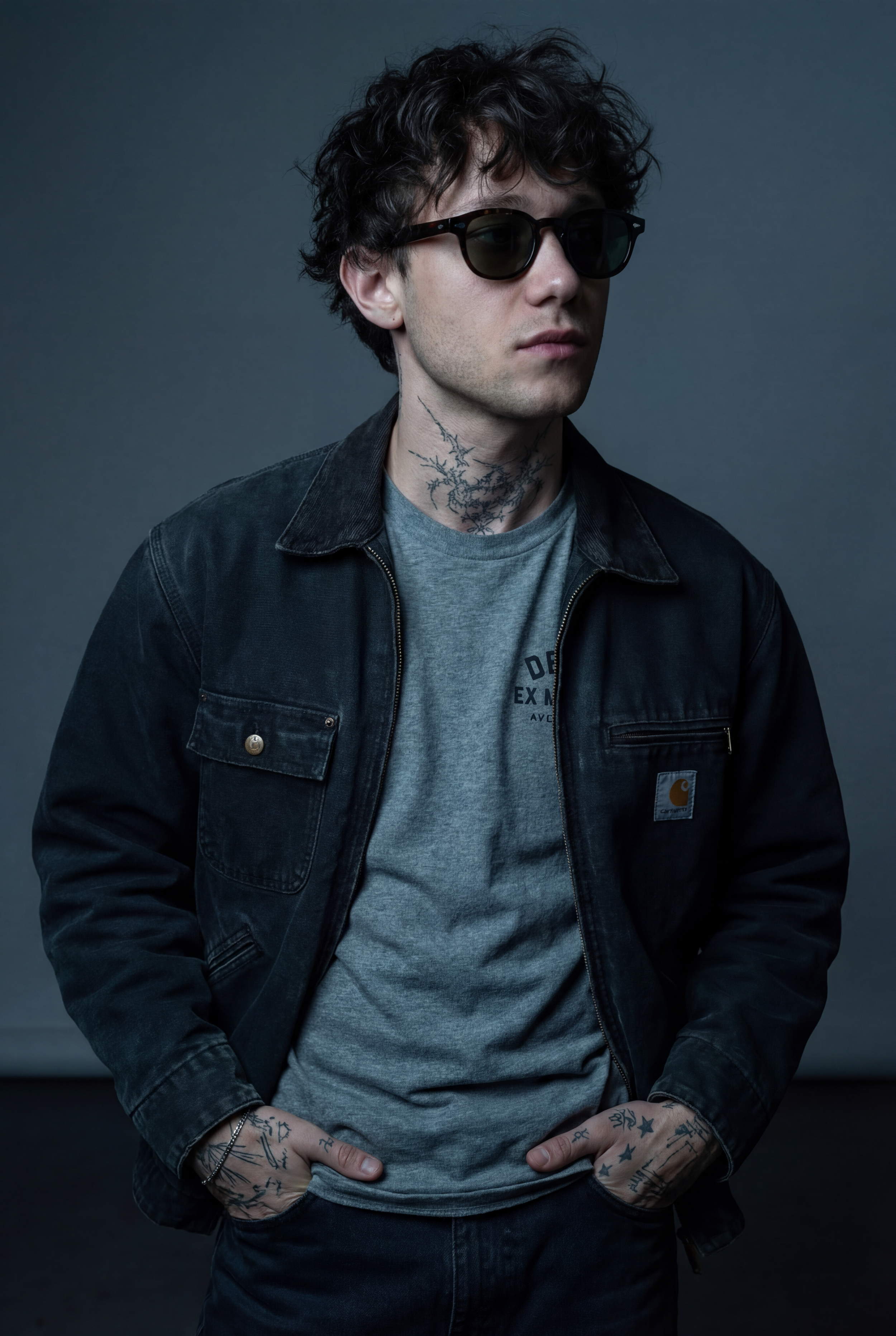 Jeune homme avec des tatouages, portant des lunettes de soleil, une veste en denim noire et un t-shirt gris, debout contre un fond gris.