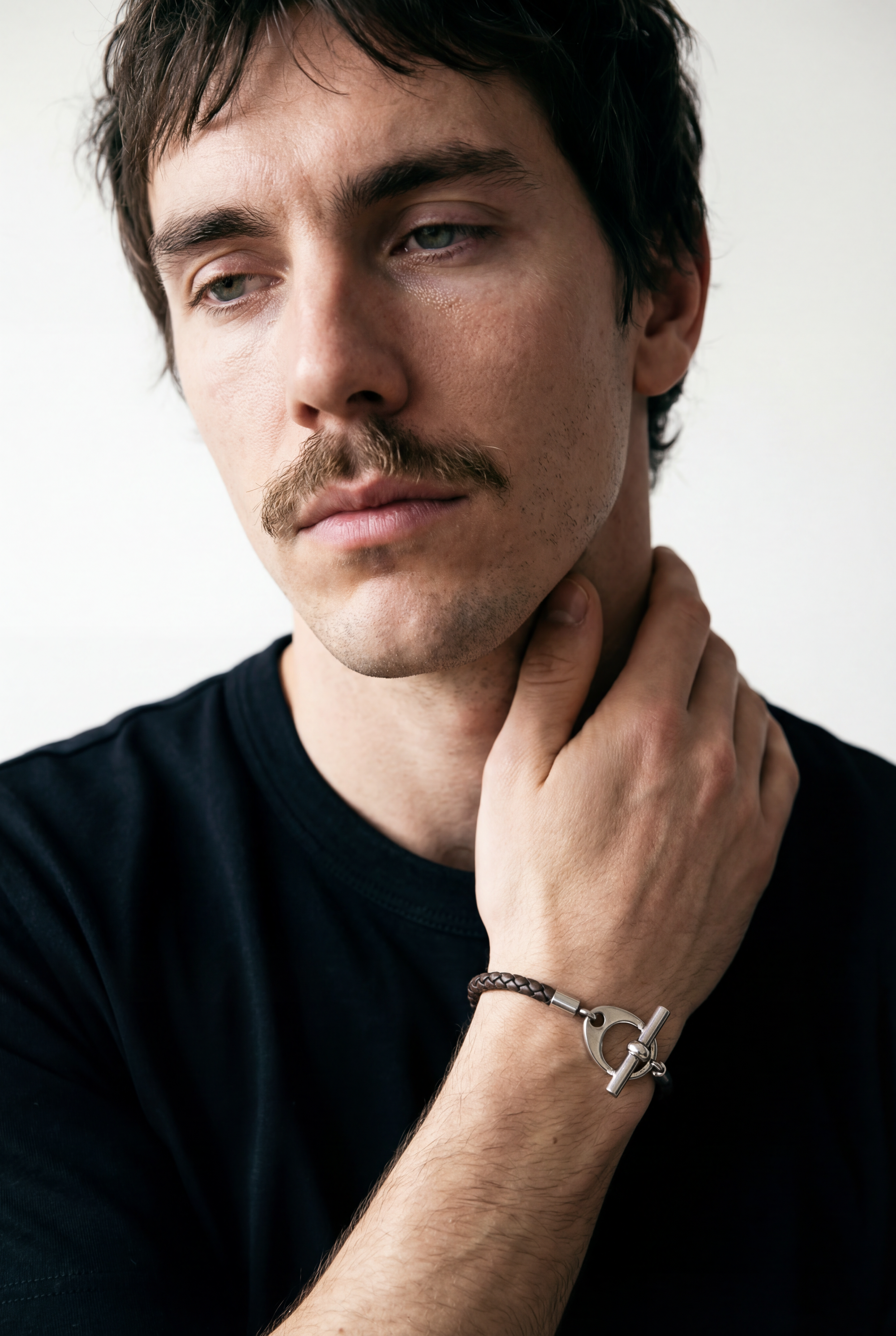 Un homme avec une moustache, portant un bracelet en cuir et un t-shirt noir, regarde pensivement, touchant sa gorge.