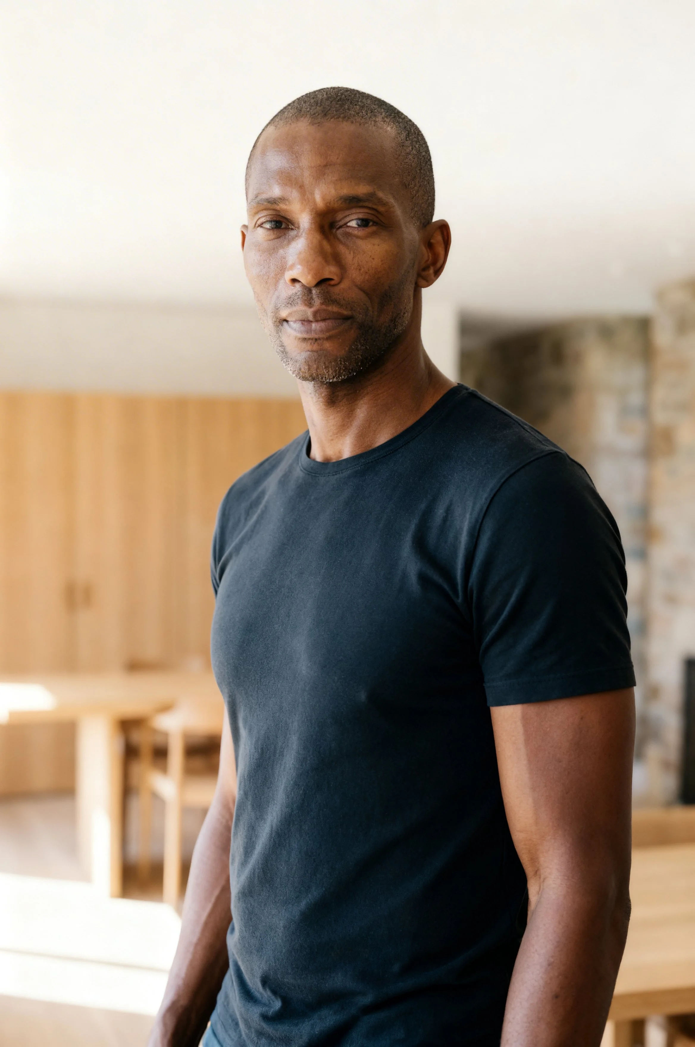 Homme debout dans une pièce lumineuse, portant un t-shirt noir, avec un arrière-plan de meubles en bois et un mur en pierre.