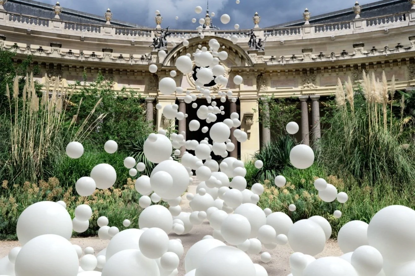 Une installation artistique composée de nombreuses sphères blanches flottant en l'air dans un jardin orné de plantes vertes et de fleurs, avec un bâtiment historique en arrière-plan, sous un ciel nuageux.
