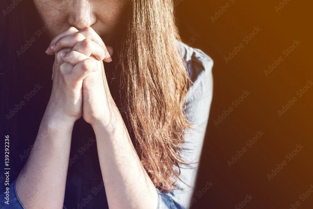 Close-up of a woman with long hair praying with her hands clasped near her face.