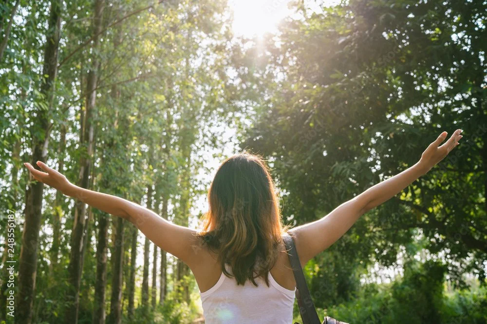 A woman with brown hair wearing a white tank top standing in a forest with her arms raised, facing the sunlight.