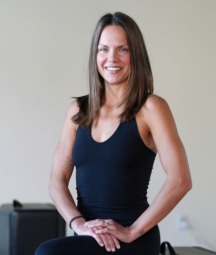 A woman with brown hair and blue eyes, smiling gently, sitting on a wooden floor with a neutral background, wearing a patterned sports bra and navy leggings, with a pearl necklace and a delicate gold necklace.