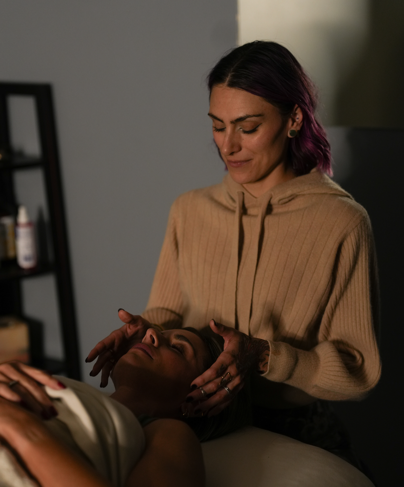 A woman with pink curly hair and tattoos is smiling and seated on a Pilates reformer machine in a fitness studio.