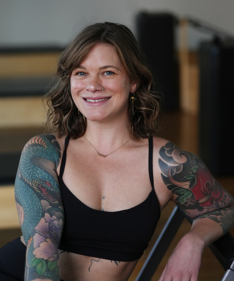A woman with long wavy blonde hair, wearing a light blue long sleeve dress, smiling and leaning against a marble wall with her arms crossed. She is outdoors with a blurred background of trees and sunlight.