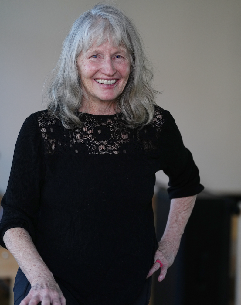 A smiling woman with gray hair and a green shirt standing in a room with natural light, hands pressed together in a prayer pose.