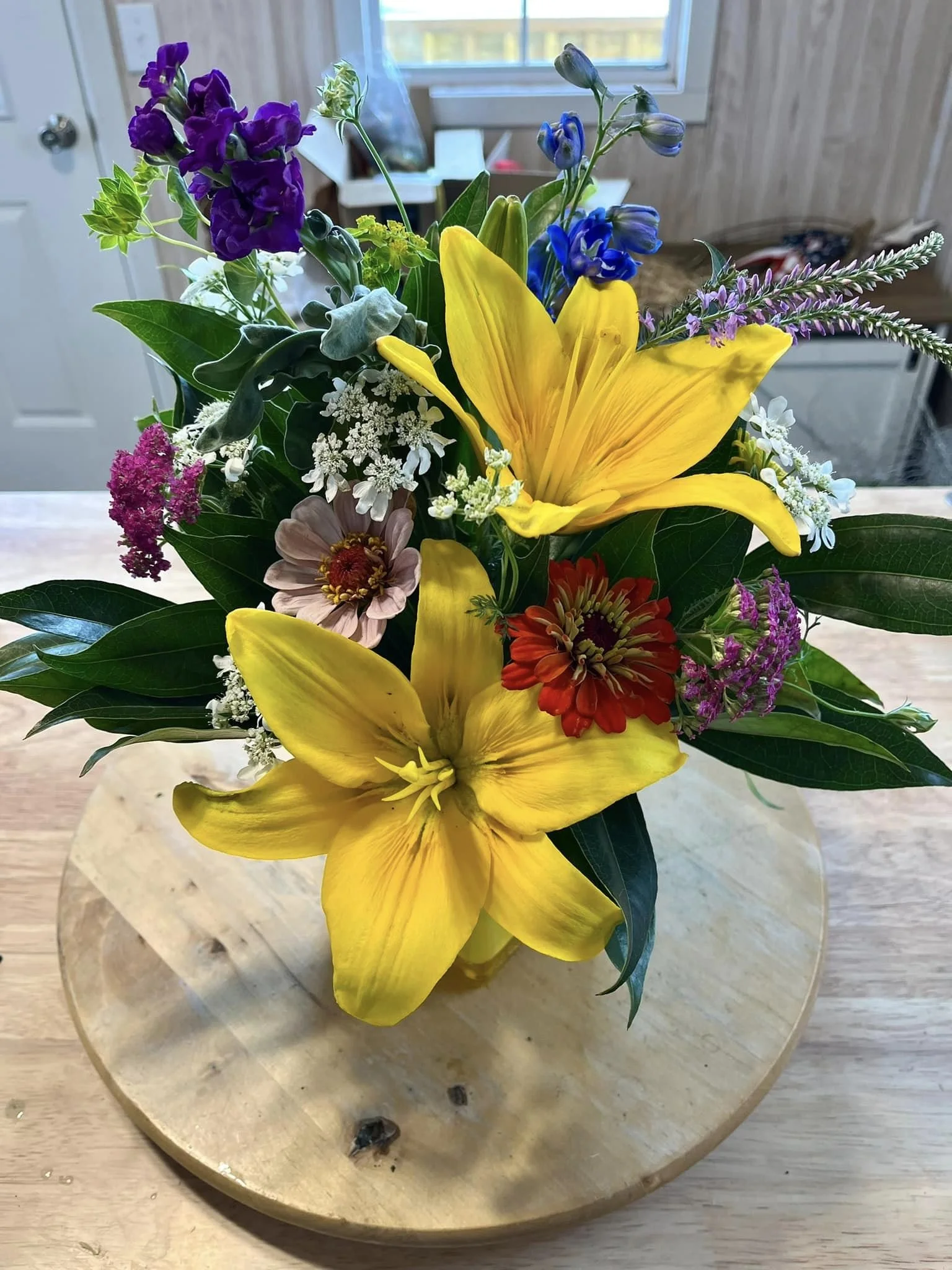 A bouquet of colorful flowers including yellow lilies, purple, white, pink, and red flowers, and green foliage in a glass vase on a wooden table.