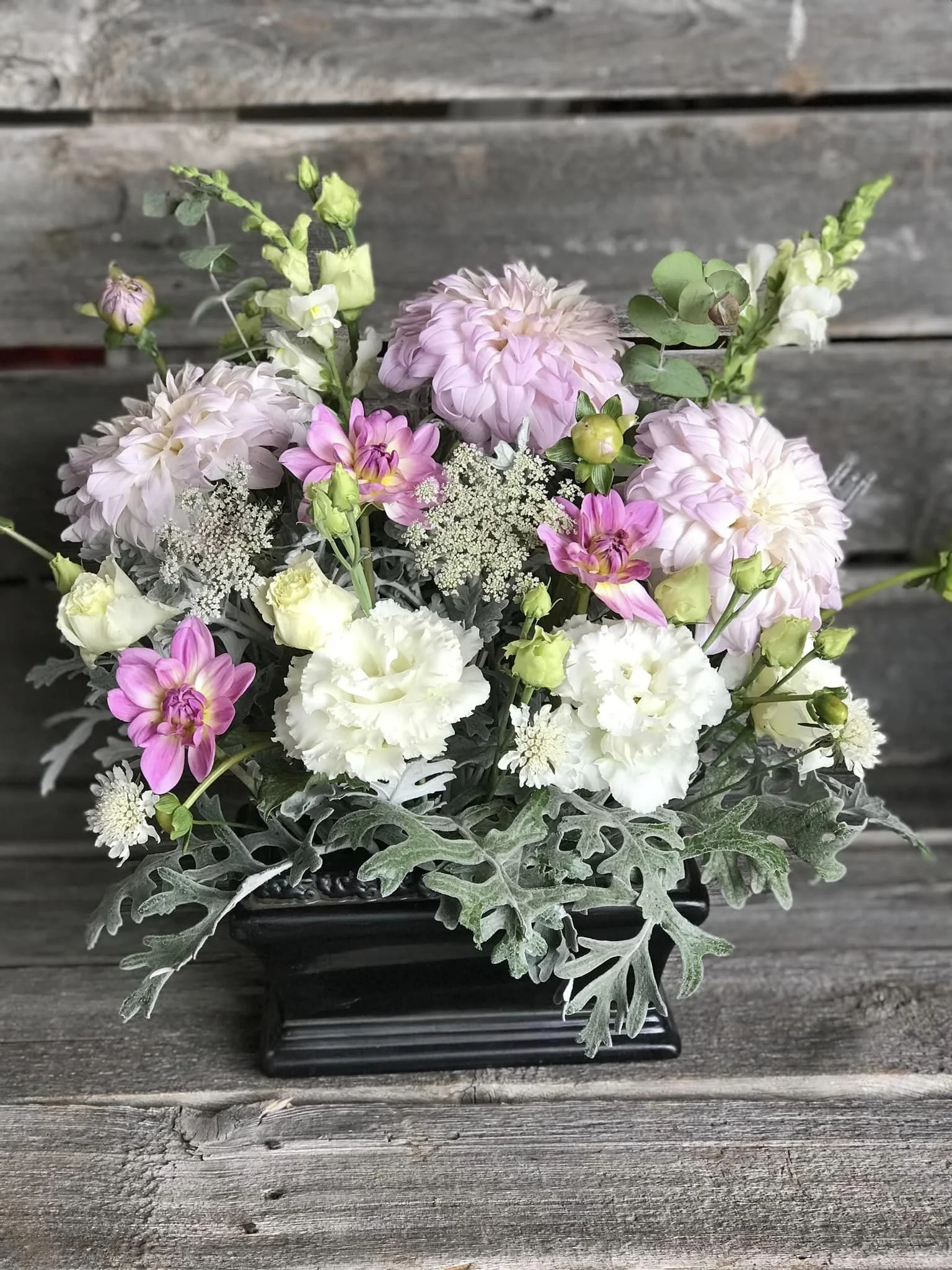 A bouquet of pink, white, and lavender flowers in a black rectangular vase on a wooden surface with a rustic wooden background.