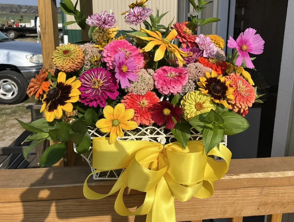 A colorful bouquet of mixed flowers in a white basket decorated with a yellow ribbon, placed on a wooden surface outdoors.