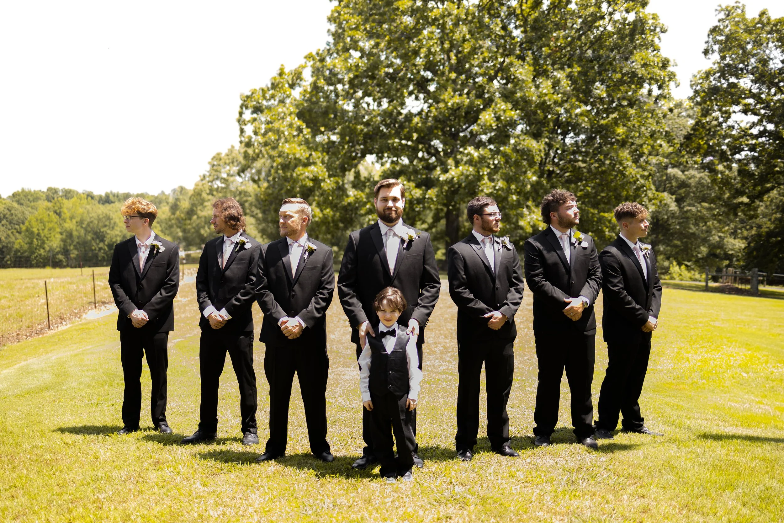 Group of seven men and one young boy in tuxedos standing on grass outdoors, with trees in the background, on a sunny day at a wedding.
