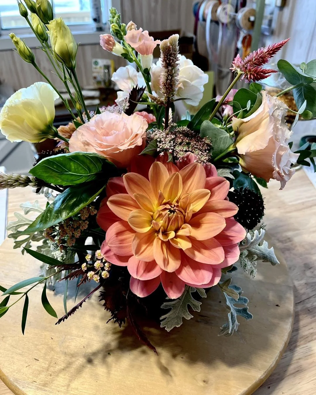 A colorful bouquet of mixed flowers including a large orange-pink dahlia, light pink roses, white lisianthus, and various greenery arranged in a vase on a wooden table.