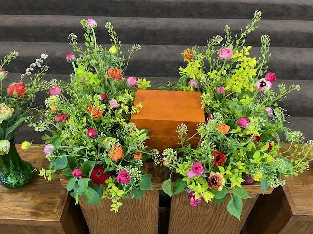 Colorful floral arrangement with various flowers surrounding a small wooden box, placed on wooden tables with a dark, textured background.