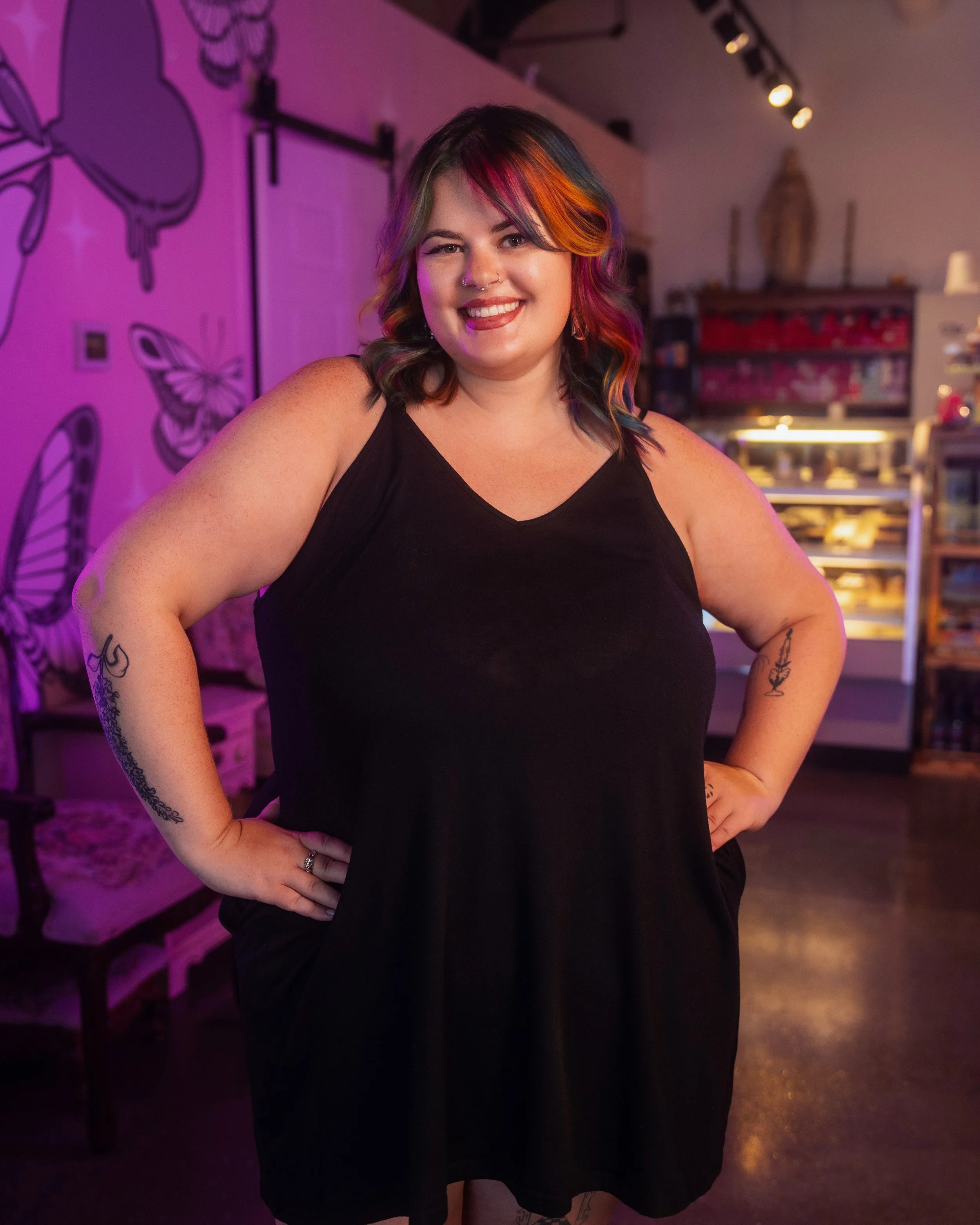 A woman with rainbow-colored hair that transitions from red at the roots to orange, yellow, green, blue, and purple toward the ends, smiling slightly, wearing a black top with a lace neckline, against a plain white background.