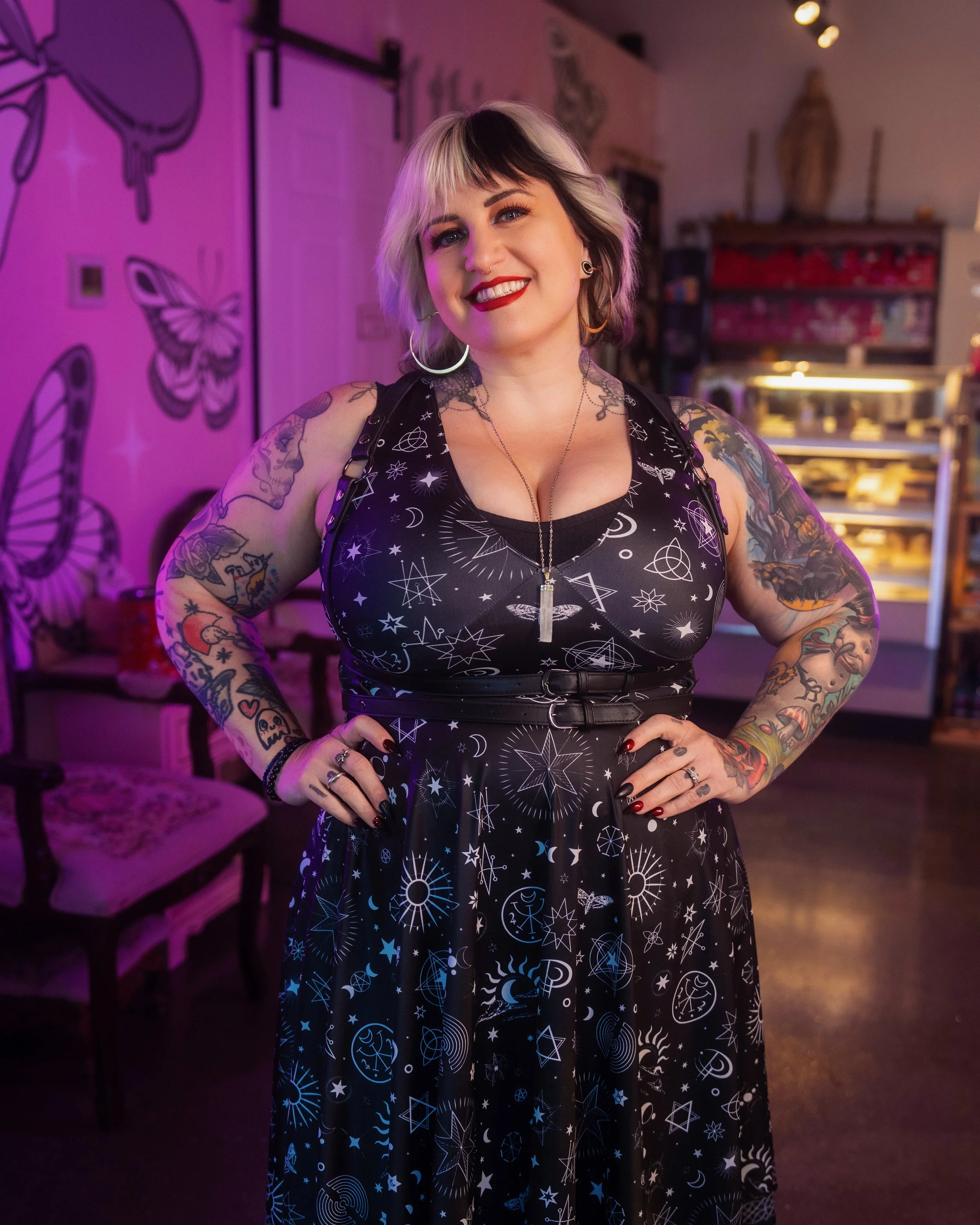 A woman with light blond hair and makeup wearing a dress with a black background and white mystical and tarot symbols, standing in front of a decorated wall with tarot and mystical themes in a store.